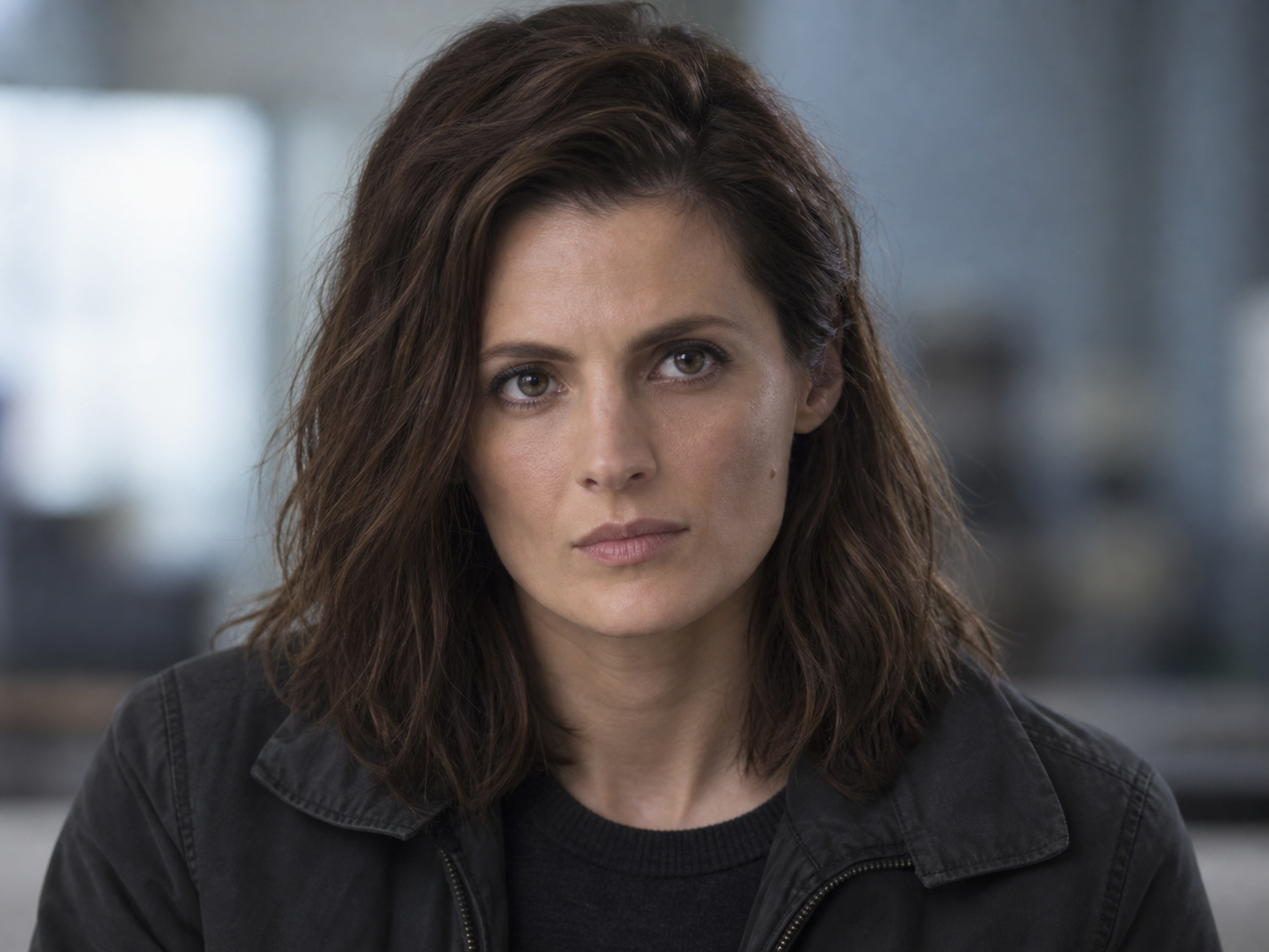 Stana Katic as Emily Byrne, with loose dark hair and a charcoal jacket against a softly blurred interior.