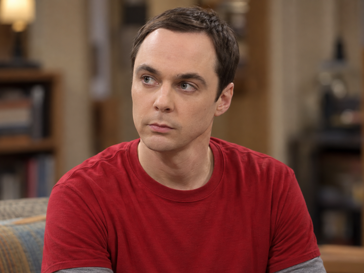 Jim Parsons as Sheldon Cooper, with short dark hair and a red T-shirt over a grey long-sleeved top, against a softly blurred interior.