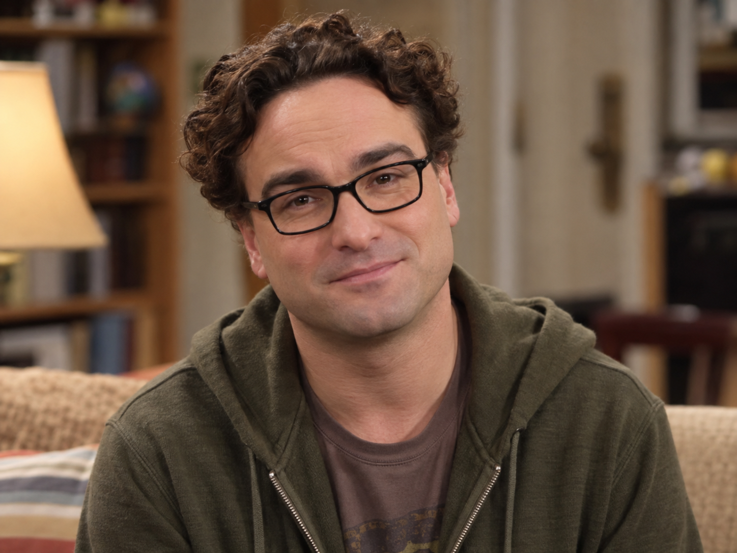 Johnny Galecki as Leonard Hofstadter, with curly brown hair, dark rectangular glasses and an olive hoodie over a muted T-shirt, against a softly blurred interior.
