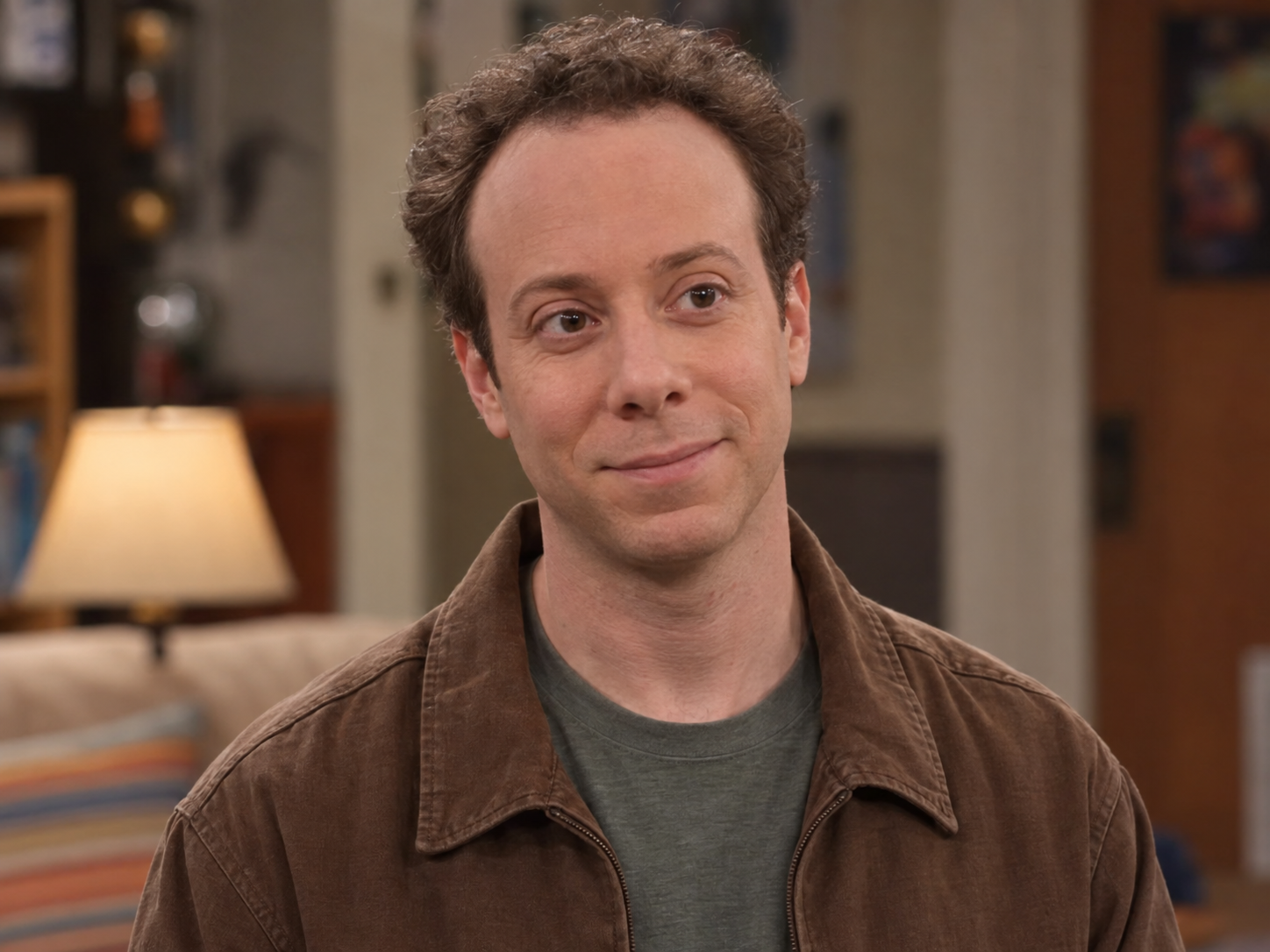 Kevin Sussman as Stuart Bloom, with short curly brown hair and a brown casual jacket over a grey T-shirt, against a softly blurred interior.