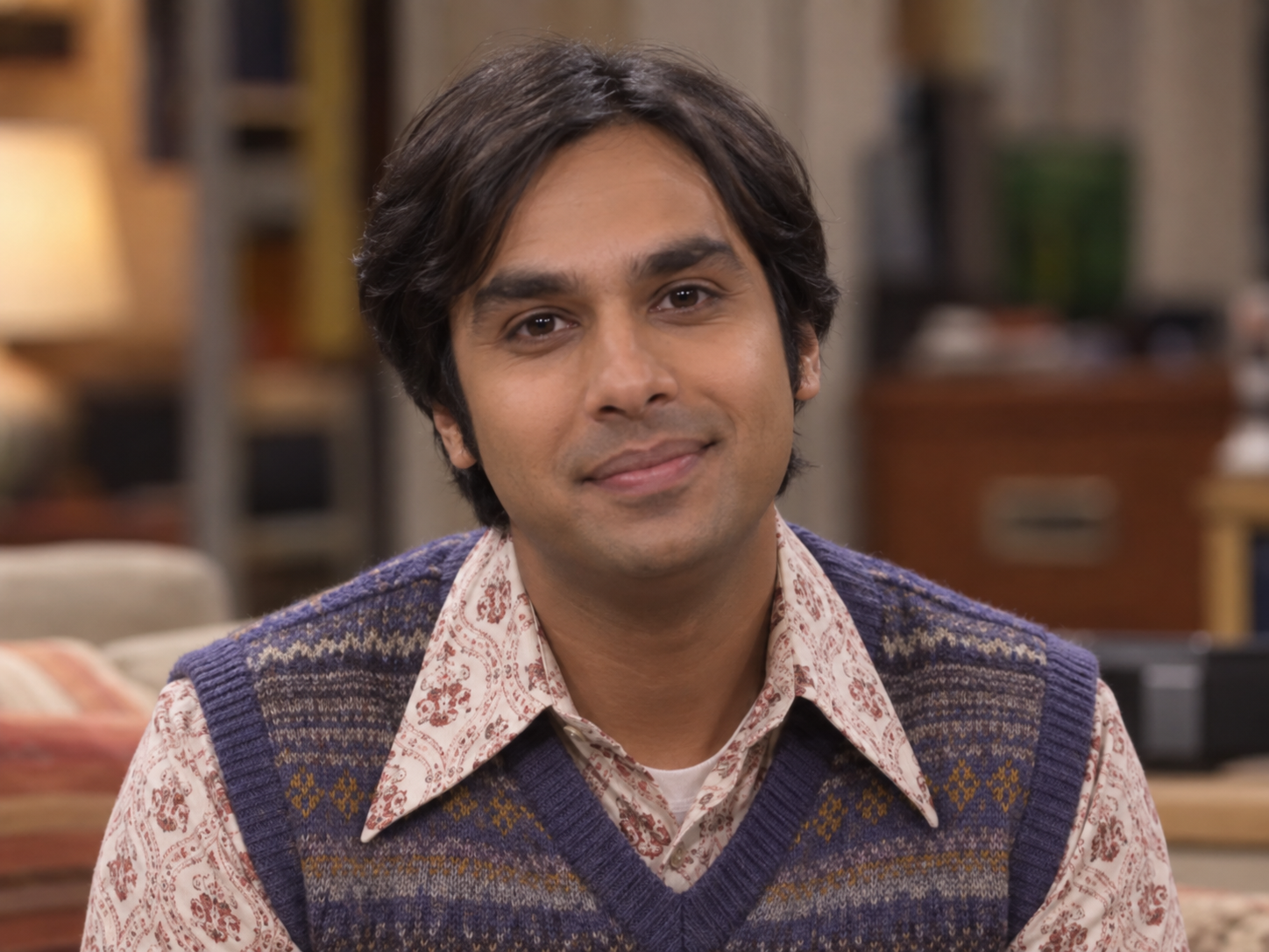 Kunal Nayyar as Raj Koothrappali, with side-parted dark hair and a blue patterned sweater vest over a paisley shirt, against a softly blurred interior.
