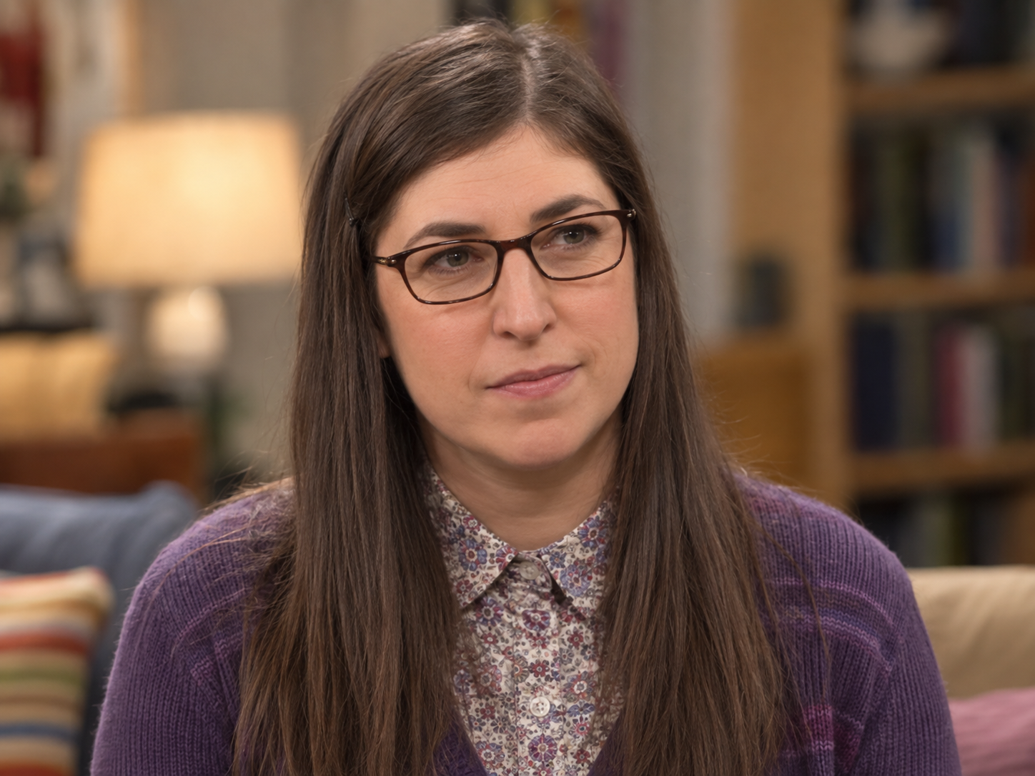 Mayim Bialik as Amy Farrah Fowler, with long straight brown hair, dark rectangular glasses and a purple cardigan over a patterned blouse, against a softly blurred interior.
