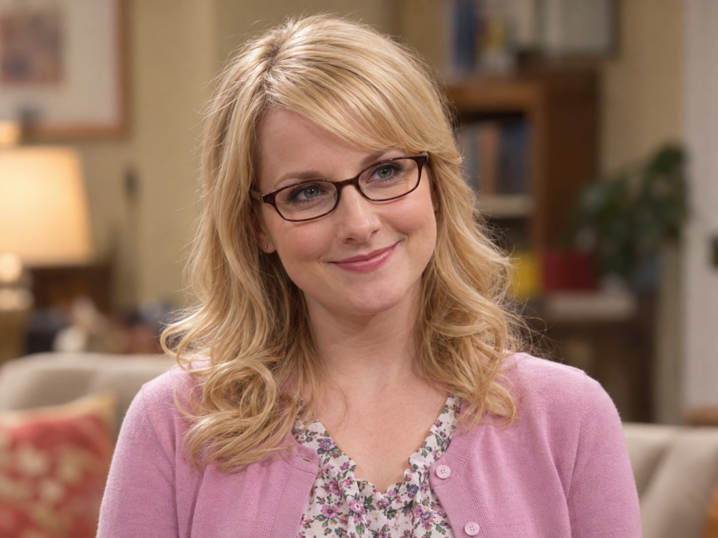 Melissa Rauch as Bernadette Rostenkowski, with blonde curls, dark-framed glasses and a pink cardigan over a floral blouse, against a softly blurred interior.