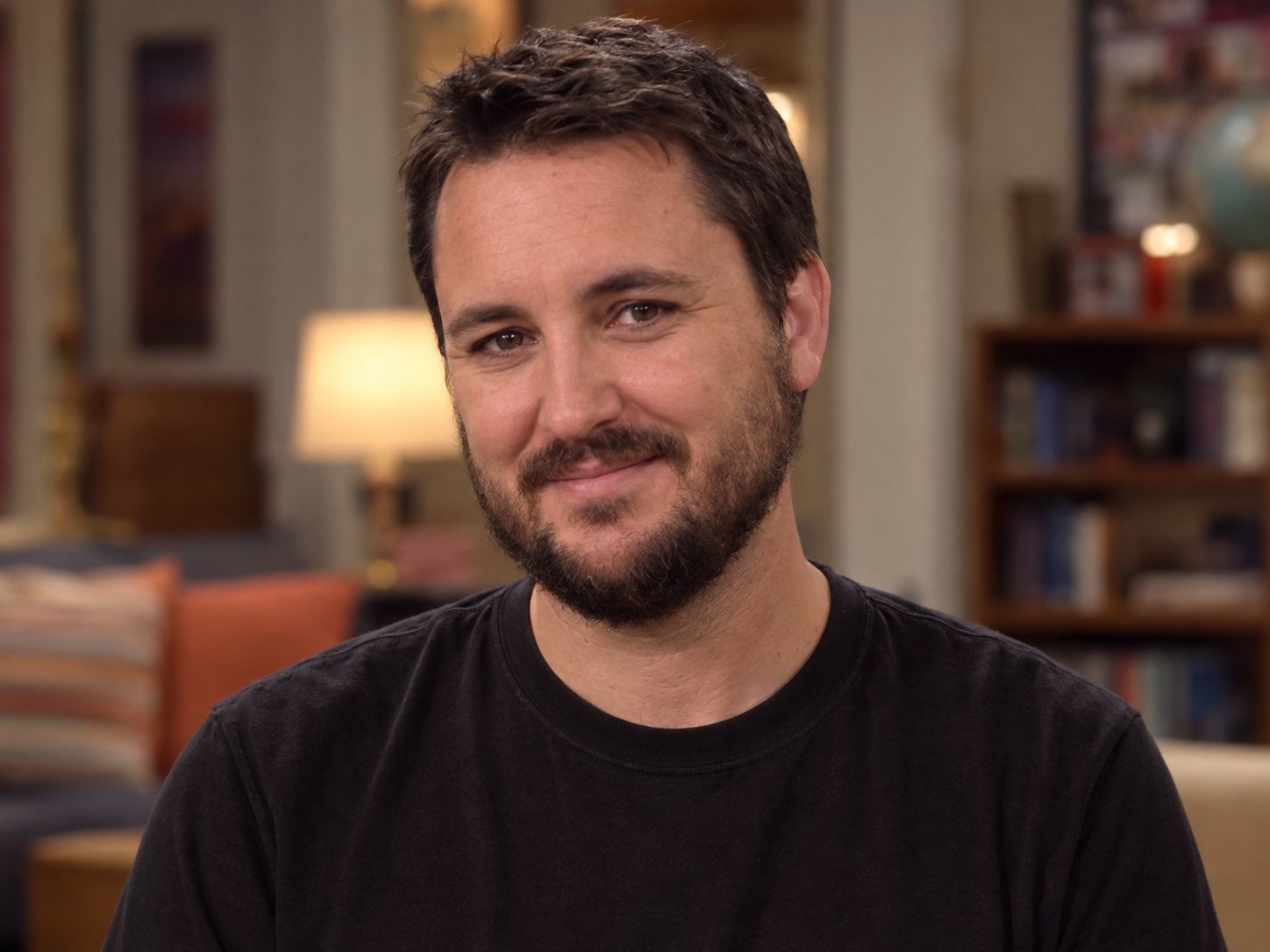 Wil Wheaton as the fictionalised Wil Wheaton, with short dark hair, a trimmed beard and a plain dark T-shirt, against a softly blurred interior.