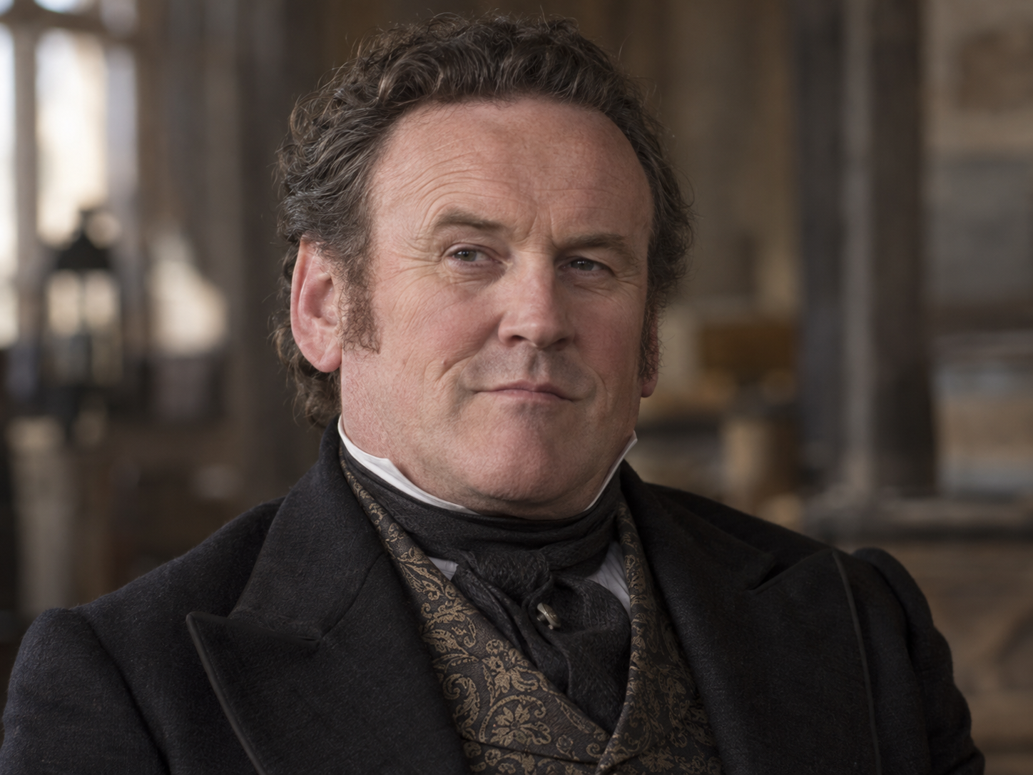Colm Meaney as Thomas Durant, in a dark coat, patterned waistcoat and cravat inside a softly lit railway carriage.