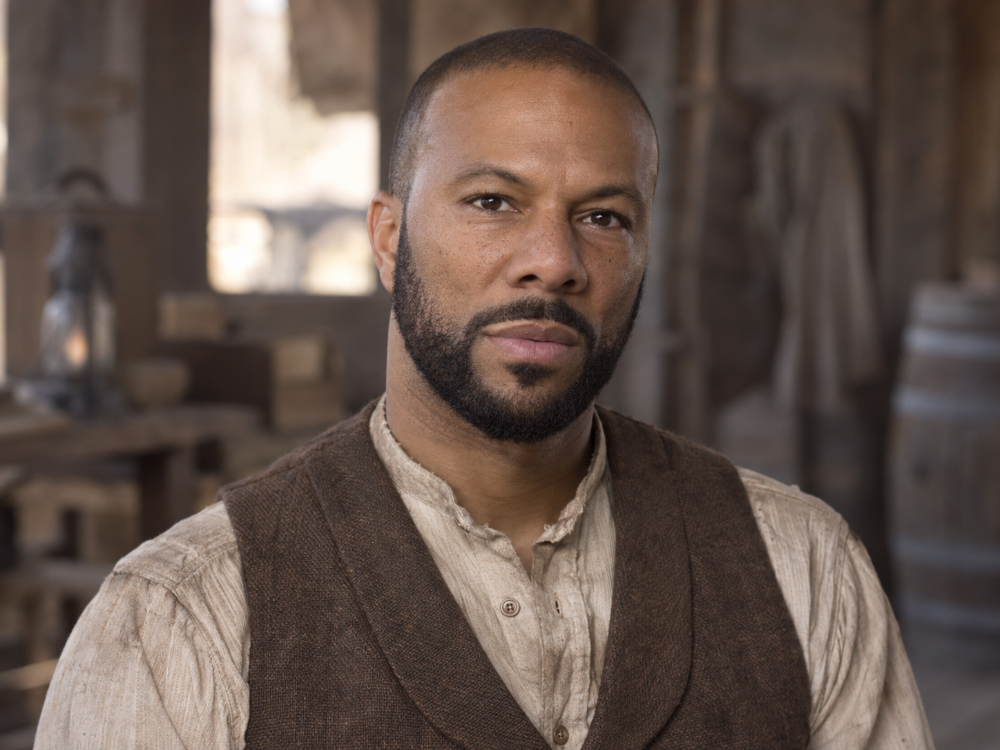 Common as Elam Ferguson, wearing a collarless shirt and brown waistcoat against a softly blurred wooden interior.