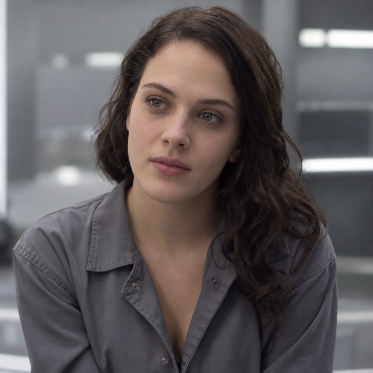 Jessica Brown Findlay as Abi, with dark hair and an open-collar grey jumpsuit, against a softly blurred futuristic interior.