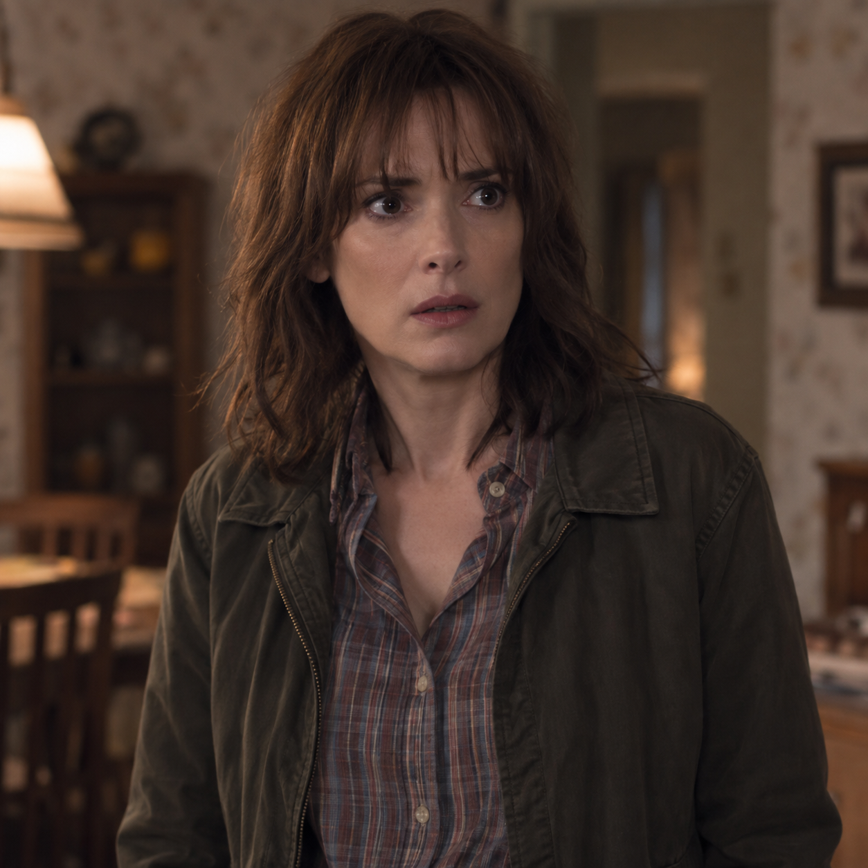 Winona Ryder as Joyce Byers, wearing a checked blouse and practical jacket in a house.